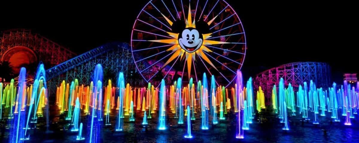 Scène nocturne colorée mettant en vedette une grande roue avec une silhouette de Mickey Mouse illuminée, entourée de luminaires vibrants et multicolores et d'autres structures éclairées.