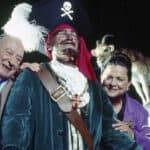 Un couple de personnes âgées posant joyeusement avec une personne habillée en pirate, avec un chapeau à plumes et un cache-œil, debout devant la statue de chèvre de Davis.