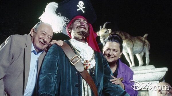 Un couple de personnes âgées posant joyeusement avec une personne habillée en pirate, avec un chapeau à plumes et un cache-œil, debout devant la statue de chèvre de Davis.