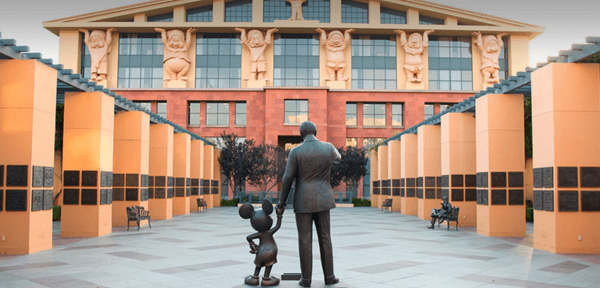Une statue de Walt Disney tenant la main de Mickey Souris, face à un grand bâtiment orné de reliefs de personnages, installé dans une cour aux escaliers symétriques.