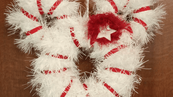 Une couronne blanche festive et moelleuse avec des rubans rayés de canne à sucre rouges et blancs et un nœud rouge foncé avec un centre étincelant, accroché contre une porte en bois.