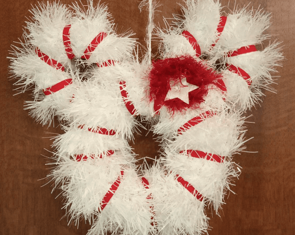 Une couronne blanche festive et moelleuse avec des rubans rayés de canne à sucre rouges et blancs et un nœud rouge foncé avec un centre étincelant, accroché contre une porte en bois.