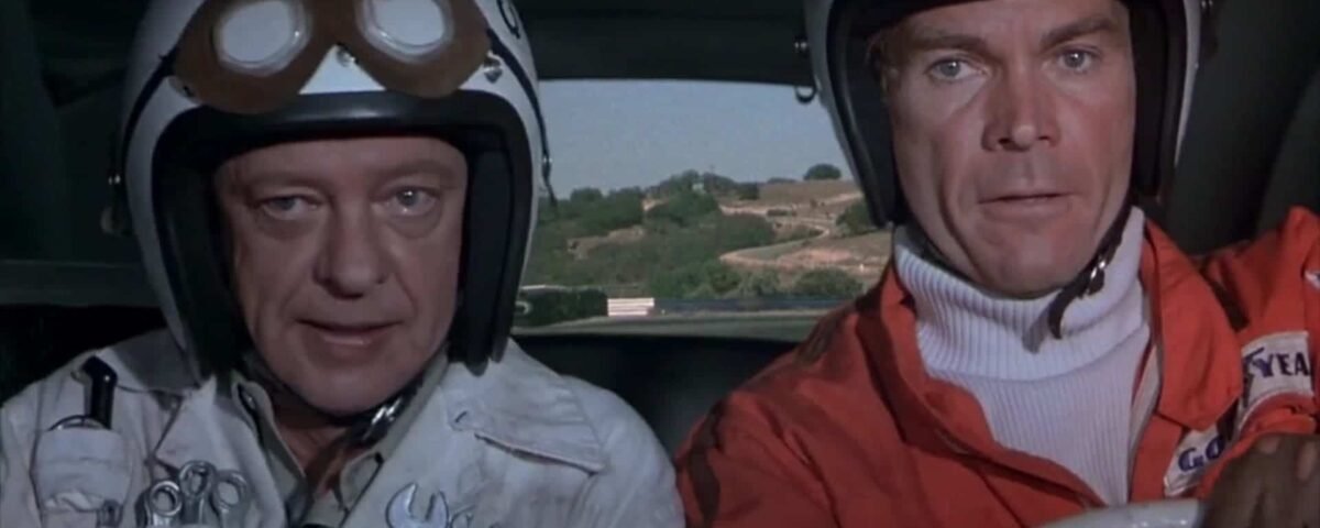Deux hommes dans une voiture portant des casques et des lunettes de course sont prêts pour "La Coccinelle à Monte Carlo". Un homme, à gauche, porte un casque blanc avec un « W » dessus.