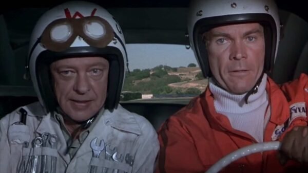 Deux hommes dans une voiture portant des casques et des lunettes de course sont prêts pour "La Coccinelle à Monte Carlo". Un homme, à gauche, porte un casque blanc avec un « W » dessus.