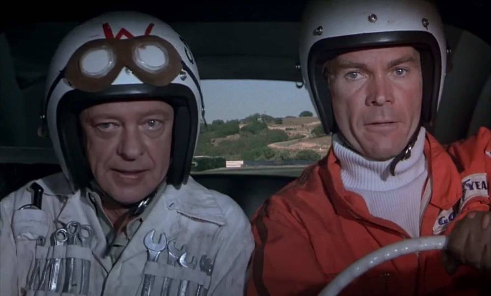 Deux hommes dans une voiture portant des casques et des lunettes de course sont prêts pour "La Coccinelle à Monte Carlo". Un homme, à gauche, porte un casque blanc avec un « W » dessus.