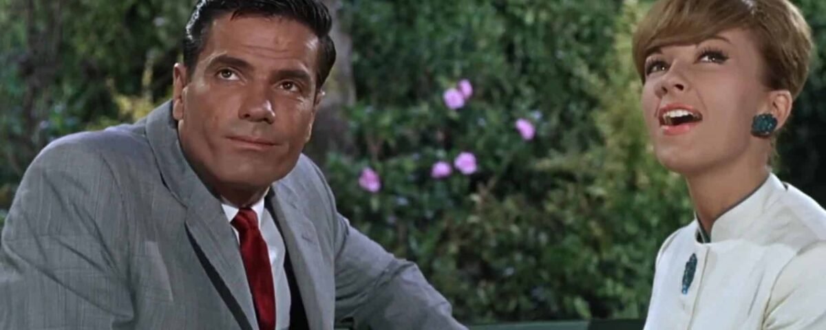 Un homme en costume gris et une femme en blouse blanche avec de grandes boucles d'oreilles vertes sont assis sur un banc, tous deux souriants alors qu'ils engagent une conversation joyeuse dans un jardin, ressemblant à un pilote.