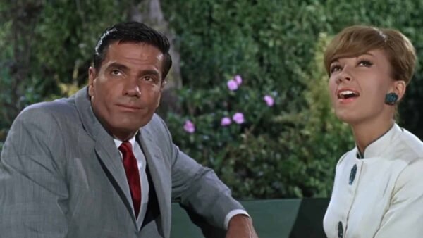 Un homme en costume gris et une femme en blouse blanche avec de grandes boucles d'oreilles vertes sont assis sur un banc, tous deux souriants alors qu'ils engagent une conversation joyeuse dans un jardin, ressemblant à un pilote.
