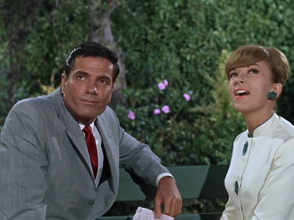 Un homme en costume gris et une femme en blouse blanche avec de grandes boucles d'oreilles vertes sont assis sur un banc, tous deux souriants alors qu'ils engagent une conversation joyeuse dans un jardin, ressemblant à un pilote.