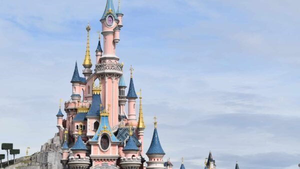 Une vue pittoresque d'un château de conte de fées avec des flèches roses et bleues sur un ciel partiellement nuageux. Ce bâtiment emblématique et fantaisiste de Disneyland Paris est entouré d'une verdure luxuriante.