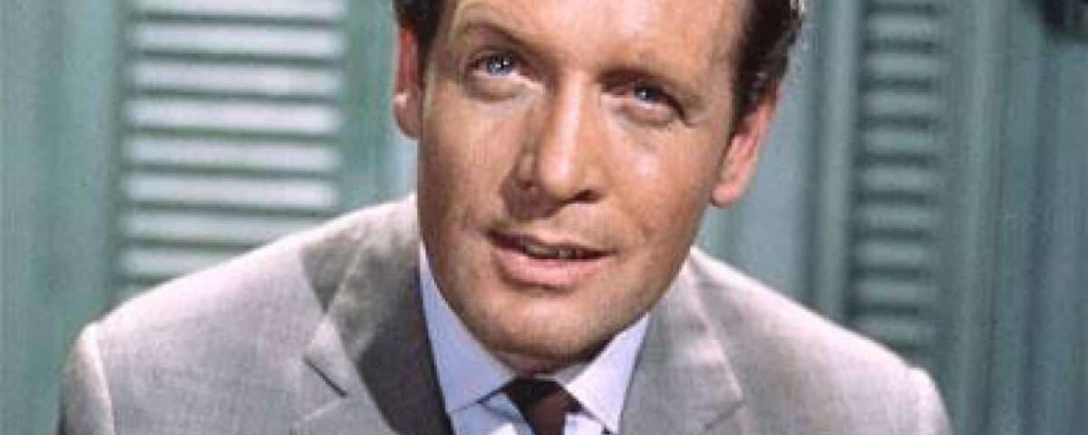 Un homme d'âge moyen aux cheveux clairs, vêtu d'un costume et d'une cravate, regarde légèrement de côté avec une expression pensive. Il est positionné devant un fond vert à lattes verticales rappelant Patrick