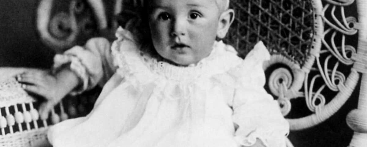 Une photo vintage en noir et blanc d'un bébé assis sur une chaise en osier conçue par Walt, vêtu d'une robe blanche avec un col en dentelle à volants. L'enfant regarde directement la caméra avec un