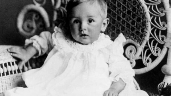 Une photo vintage en noir et blanc d'un bébé assis sur une chaise en osier conçue par Walt, vêtu d'une robe blanche avec un col en dentelle à volants. L'enfant regarde directement la caméra avec un