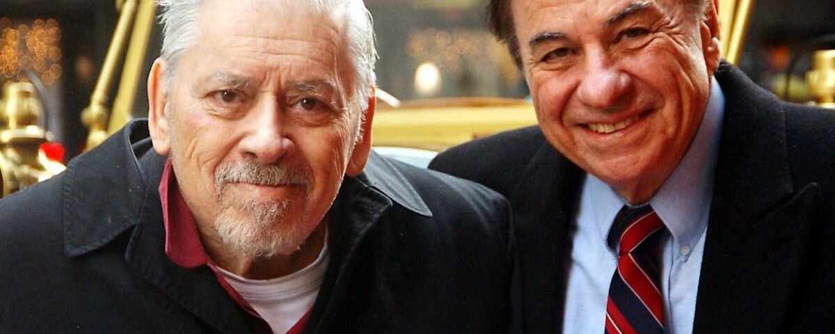 Deux hommes âgés souriants, l'un avec des cheveux blancs et une barbe rappelant un personnage d'Alice au pays des merveilles, l'autre avec des cheveux noirs et une cravate, rapprochés lors d'un événement.