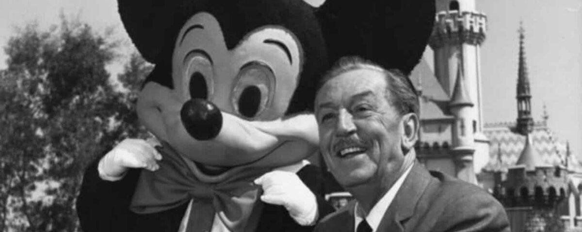 Walt Disney souriant aux côtés de Mickey Souris à Disneyland, avec le château emblématique du parc en arrière-plan.