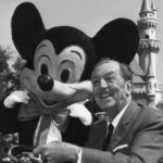 Walt Disney souriant aux côtés de Mickey Souris à Disneyland, avec le château emblématique du parc en arrière-plan.