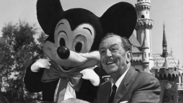 Walt Disney souriant aux côtés de Mickey Souris à Disneyland, avec le château emblématique du parc en arrière-plan.