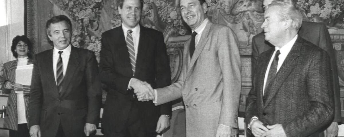 Quatre hommes en costume se serrant la main lors d'un événement officiel à Euro Disneyland, avec des tapisseries complexes en arrière-plan, peut-être dans un cadre gouvernemental ou institutionnel. Deux personnes supplémentaires sont partiellement visibles en arrière-plan