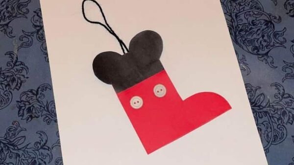 Une décoration de sapin de Noël en papier fabriquée à la main en forme de bas Mickey, avec un haut noir et un bas rouge, ornée de deux détails de boutons blancs et d'une boucle noire pour accrocher, placée sur un motif bleu.