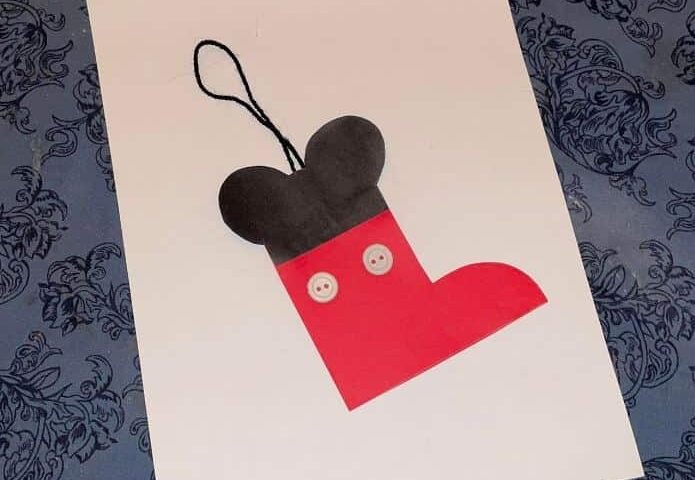 Une décoration de sapin de Noël en papier fabriquée à la main en forme de bas Mickey, avec un haut noir et un bas rouge, ornée de deux détails de boutons blancs et d'une boucle noire pour accrocher, placée sur un motif bleu.