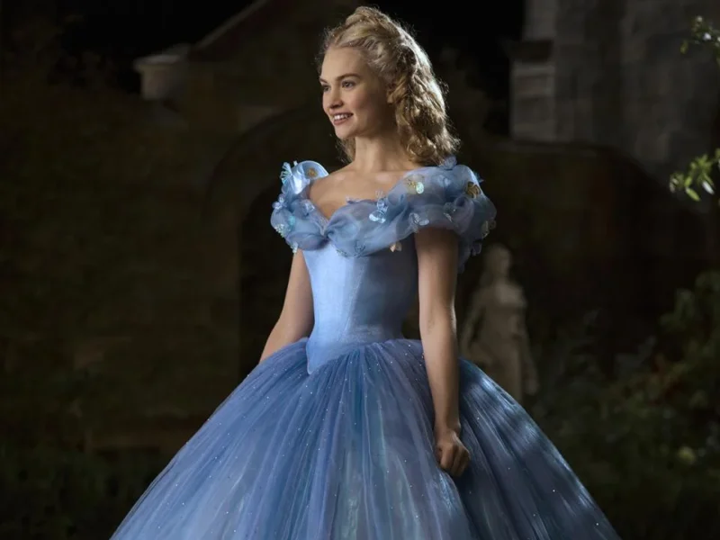 Une femme habillée en Cendrillon se tient dans une grande cour. Elle porte une élégante robe bleue avec de délicats accents floraux et ses cheveux sont coiffés d'un chignon orné. Le décor est la nuit,