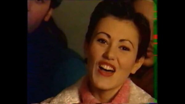 Une femme aux cheveux noirs coiffée d'un chignon, portant une écharpe et un chemisier roses, souriant joyeusement lors d'un événement Disney, avec des visages flous d'autres personnes en arrière-plan.