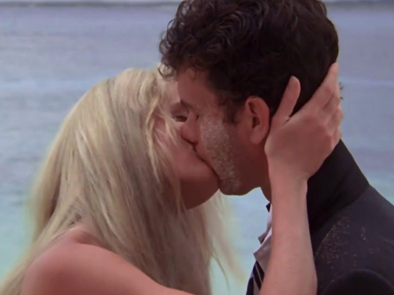 Un homme en costume noir et une femme aux cheveux blonds s'embrassent passionnément sur une plage, avec des eaux bleues calmes faisant de douces éclaboussures en arrière-plan.