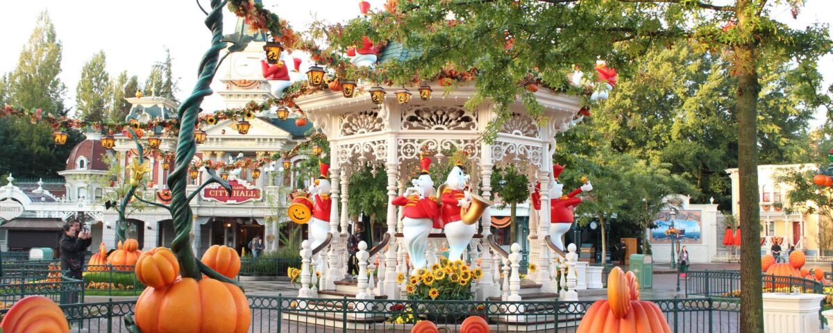 Un carrousel festif décoré d'ornements sur le thème d'Halloween, notamment de grandes citrouilles et des personnages fantômes, situé à Disneyland au milieu de plantes et d'arbres en fleurs sous un ciel dégagé.