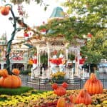 Un carrousel festif décoré d'ornements sur le thème d'Halloween, notamment de grandes citrouilles et des personnages fantômes, situé à Disneyland au milieu de plantes et d'arbres en fleurs sous un ciel dégagé.