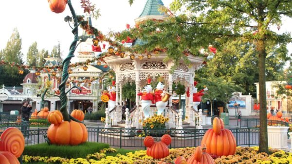 Un carrousel festif décoré d'ornements sur le thème d'Halloween, notamment de grandes citrouilles et des personnages fantômes, situé à Disneyland au milieu de plantes et d'arbres en fleurs sous un ciel dégagé.
