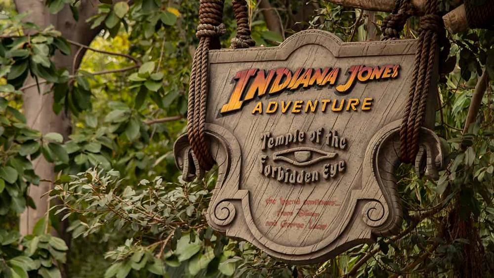 Un panneau en bois patiné pour l'attraction « Indiana Jones Adventure : Temple of the Forbidden Eye », suspendu à d'épaisses cordes sur fond de feuillage vert dense.