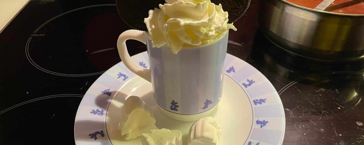 Un chocolat chaud fantaisiste garni de crème fouettée et d'un biscuit Oreo en forme de Mickey, servi sur une assiette avec des guimauves, accompagné d'un pot de chocolat chaud en fond.