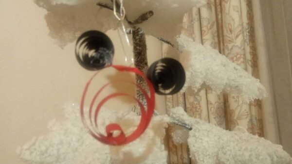 Gros plan d'un ornement Mickey fantaisiste fabriqué à la main avec un cadre rouge circulaire avec des détails en spirale et deux perles noires suspendues près du sommet, sur un tissu décoratif doux et blanc en arrière-plan.