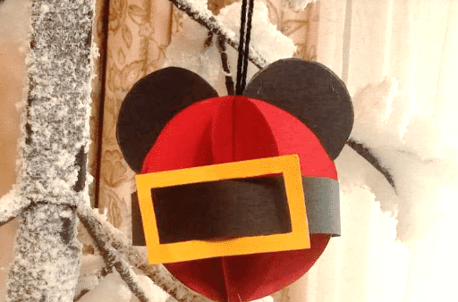 Un ornement festif ressemblant à Mickey Mouse en rouge et noir est suspendu à une branche d'arbre saupoudrée de neige. L'ornement présente un carré jaune ressemblant à une poche devant.