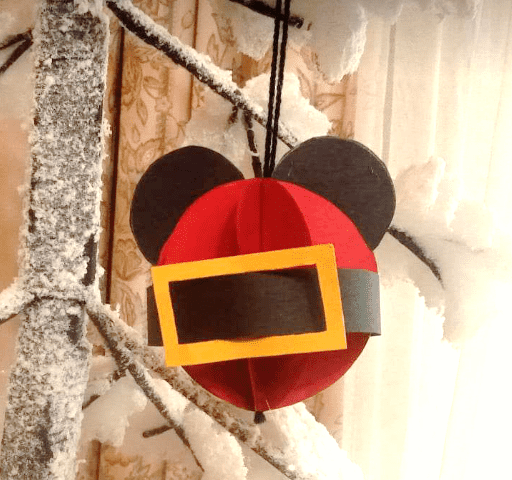 Un ornement festif ressemblant à Mickey Mouse en rouge et noir est suspendu à une branche d'arbre saupoudrée de neige. L'ornement présente un carré jaune ressemblant à une poche devant.