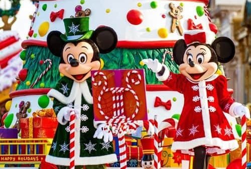 Mickey et Minnie Mouse, vêtus de tenues de vacances, se tiennent devant un gâteau de Noël festif à plusieurs étages décoré d'ornements et de cadeaux, dans un cadre lumineux et joyeux.