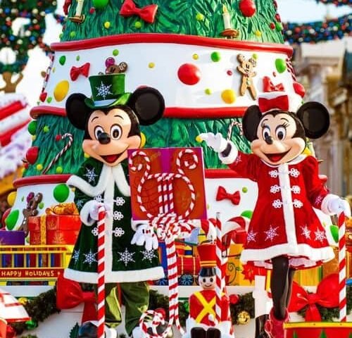 Mickey et Minnie Mouse, vêtus de tenues de vacances, se tiennent devant un gâteau de Noël festif à plusieurs étages décoré d'ornements et de cadeaux, dans un cadre lumineux et joyeux.
