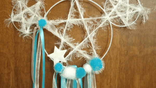 Attrape-rêves fait main avec trois anneaux interconnectés, décoré de rubans bleus et blancs, de boules moelleuses et d'une étoile en forme de Mickey, suspendu sur un fond en bois.