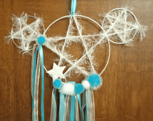 Attrape-rêves fait main avec trois anneaux interconnectés, décoré de rubans bleus et blancs, de boules moelleuses et d'une étoile en forme de Mickey, suspendu sur un fond en bois.
