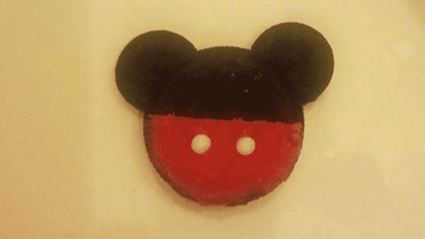 Un biscuit en forme de Mickey Mouse avec des oreilles et un dessus noirs, et un dessous rouge avec deux points blancs, ressemblant à des boutons, posés sur une assiette blanche.