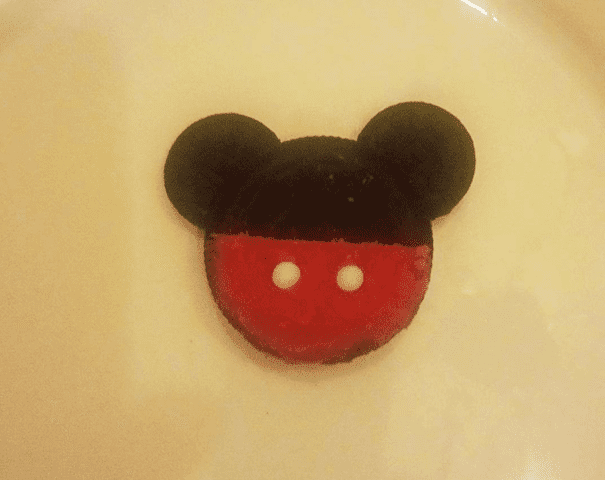 Un biscuit en forme de Mickey Mouse avec des oreilles et un dessus noirs, et un dessous rouge avec deux points blancs, ressemblant à des boutons, posés sur une assiette blanche.