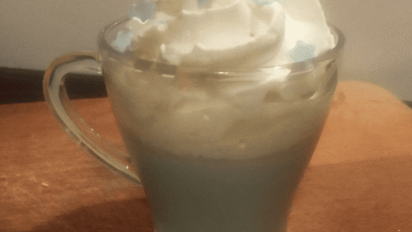 Une tasse de café en verre garnie de crème fouettée et parsemée d'étoiles comestibles bleues et de copeaux de chocolat, posée sur une table en bois sous un éclairage chaleureux.