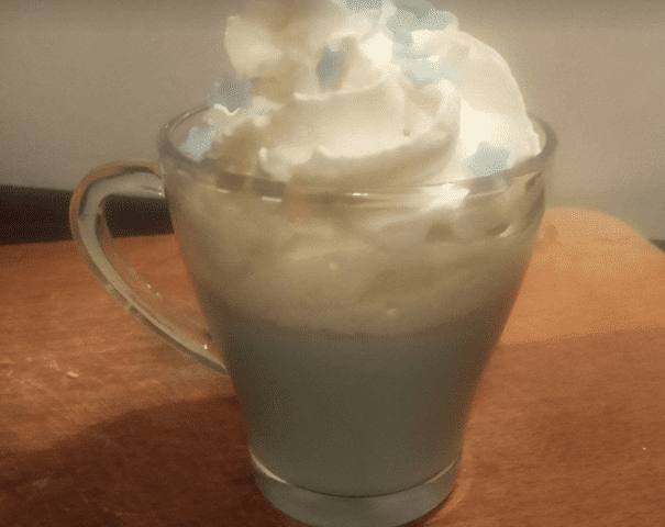 Une tasse de café en verre garnie de crème fouettée et parsemée d'étoiles comestibles bleues et de copeaux de chocolat, posée sur une table en bois sous un éclairage chaleureux.