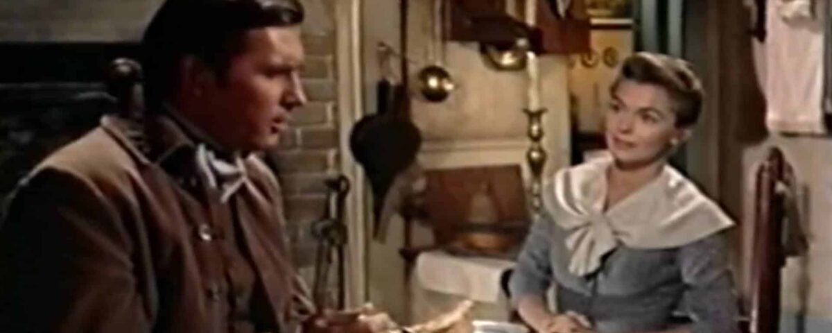 Un homme et une femme vêtus de vêtements du XIXe siècle sont assis à une table dans une cuisine rustique et discutent. L'homme tient un morceau de pain, tandis que la femme écoute attentivement :