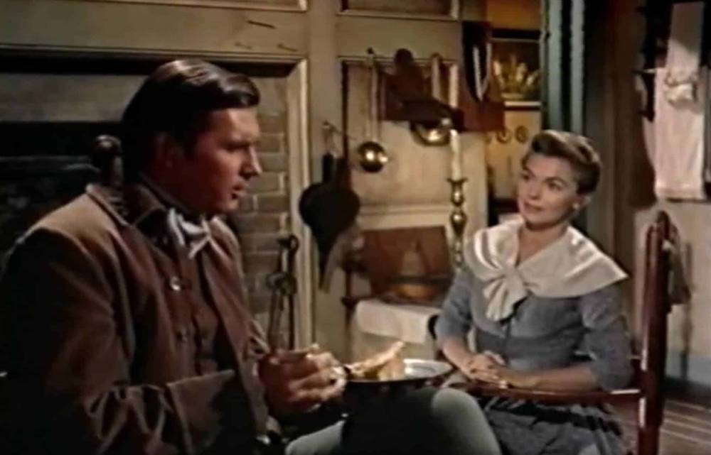 Un homme et une femme vêtus de vêtements du XIXe siècle sont assis à une table dans une cuisine rustique et discutent. L'homme tient un morceau de pain, tandis que la femme écoute attentivement :