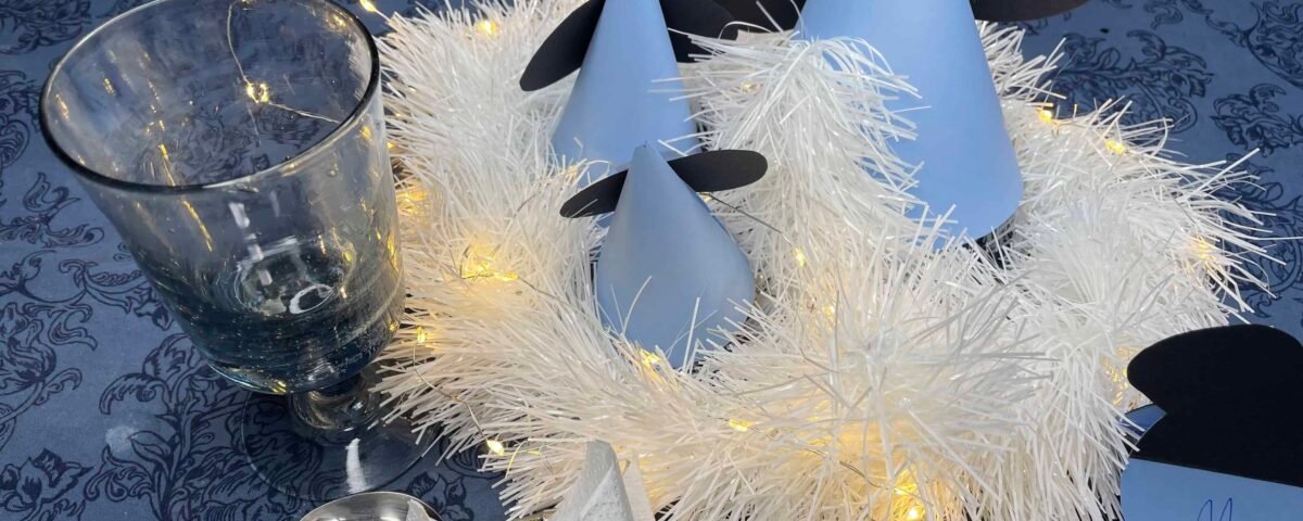 Une table de fête comprenant une pièce maîtresse avec des lumières blanches brillantes entrelacées dans une guirlande blanche moelleuse, flanquée de décorations en papier en forme de cône bleu et gris sur le thème de Mickey et d'un verre transparent.