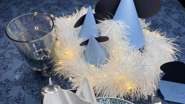 Une table de fête comprenant une pièce maîtresse avec des lumières blanches brillantes entrelacées dans une guirlande blanche moelleuse, flanquée de décorations en papier en forme de cône bleu et gris sur le thème de Mickey et d'un verre transparent.