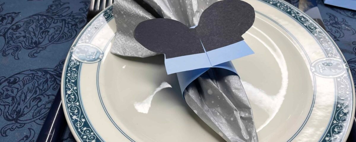 Une table de fête avec une assiette blanche à bordure bleue, une serviette argentée pliée dans un design élégant avec une découpe de papier noir en forme de Mickey et un ruban bleu dessus. Il y a des ustensiles