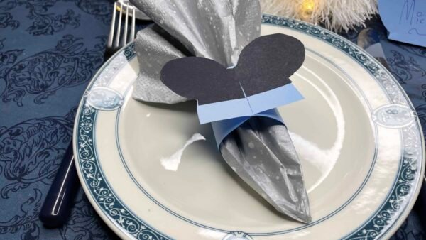 Une table de fête avec une assiette blanche à bordure bleue, une serviette argentée pliée dans un design élégant avec une découpe de papier noir en forme de Mickey et un ruban bleu dessus. Il y a des ustensiles