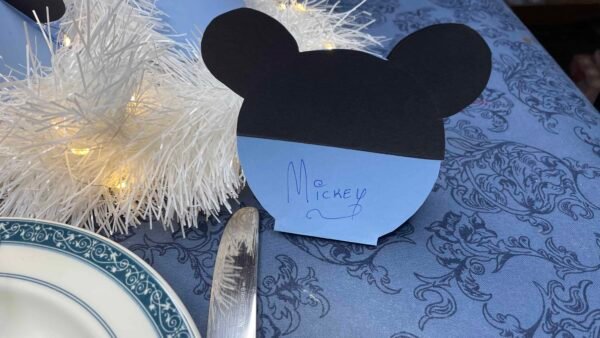 Un espace réservé en papier en forme de Mickey Mouse avec l'inscription « Mickey » dessus, dans une salle à manger avec un sapin de Noël blanc et une nappe bleue.