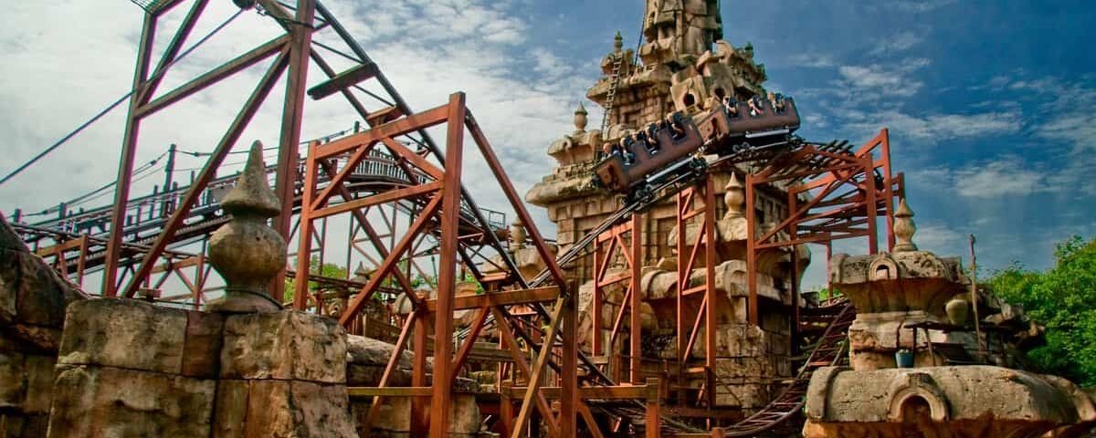 Une image d'une montagne russe avec un design de piste complexe, se faufilant autour et à travers une structure ornée en forme de tour ressemblant à des ruines antiques d'une aventure d'Indiana Jones, sur un ciel bleu avec des nuages.